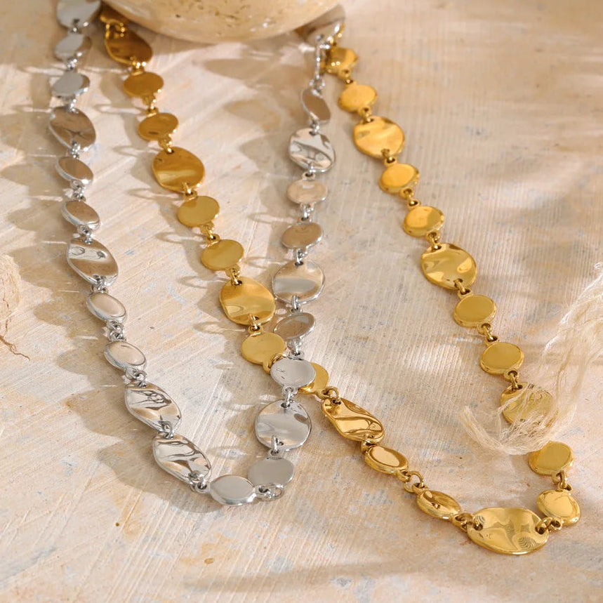 Two beaded necklaces with gold and silver disc-like beads, arranged on a textured beige fabric next to a decorative shell.