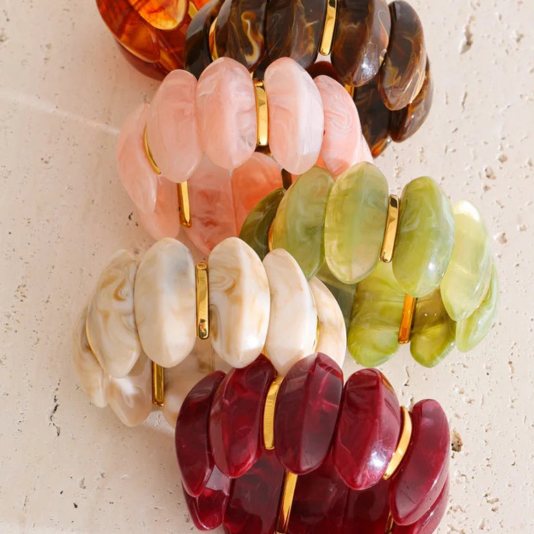 A set of six chunky disc-style bracelets with gold-tone metal accents, arranged diagonally on a light beige textured surface. The bracelets come in amber, dark brown, light pink, light green, cream, and deep red translucent colors.
