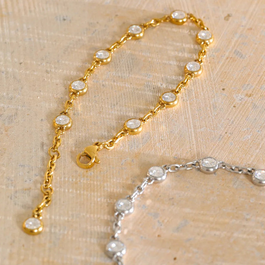 A gold chain bracelet with small clear round stones set in gold bezels, shown on a textured beige surface. A second matching bracelet in silver is partially visible in the lower right corner.