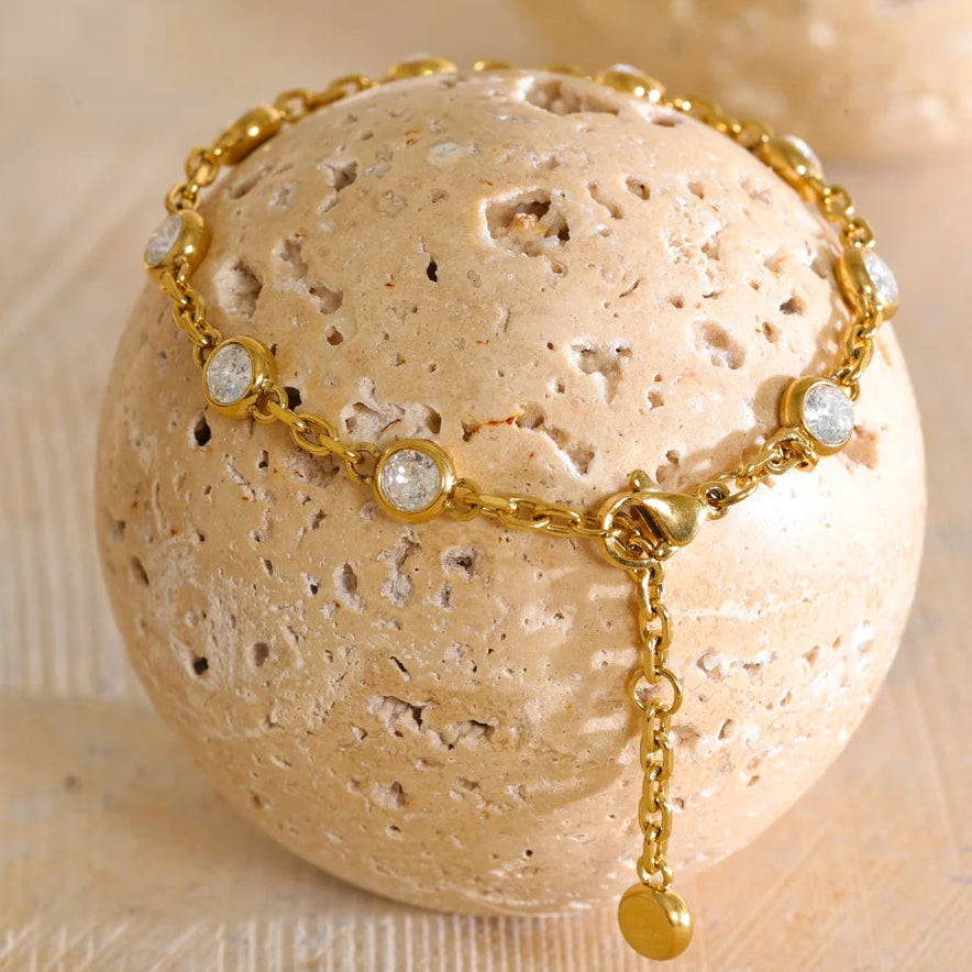 A gold chain bracelet with clear round gemstones and a dangling charm, displayed on a textured beige stone sphere against a light neutral background.