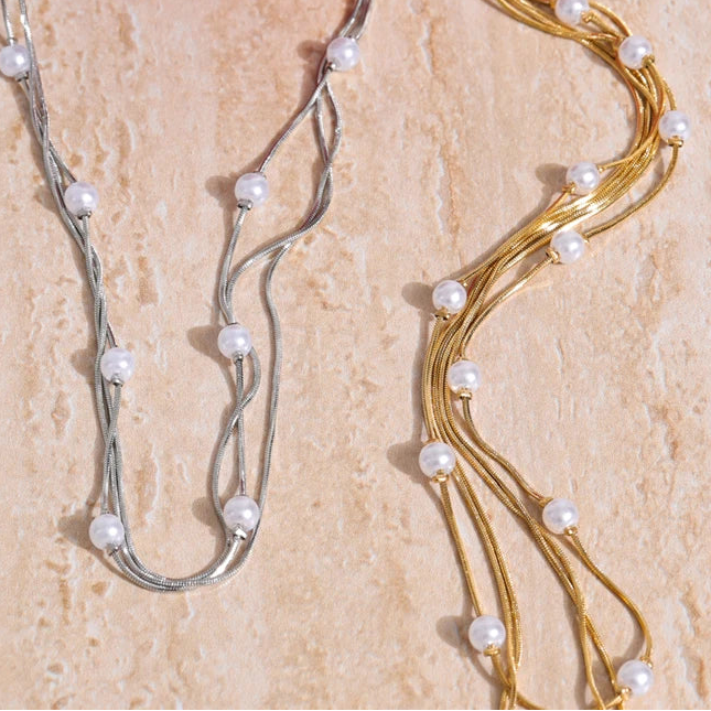 Two necklaces, one silver and one gold, with pearls on a beige surface next to an apple.