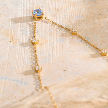 A gold-toned dangle necklace featuring a round light blue gemstone at the top and four smaller clear stones along the chain, displayed on a textured beige surface.