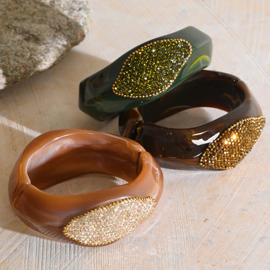Three chunky resin bangles with leaf-shaped crystal embellishments, displayed on a light wood surface. The foreground bangle is a warm brown with a gold-toned crystal leaf.