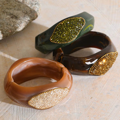 Three chunky resin bangles with leaf-shaped crystal embellishments, displayed on a light wood surface. The foreground bangle is a warm brown with a gold-toned crystal leaf.
