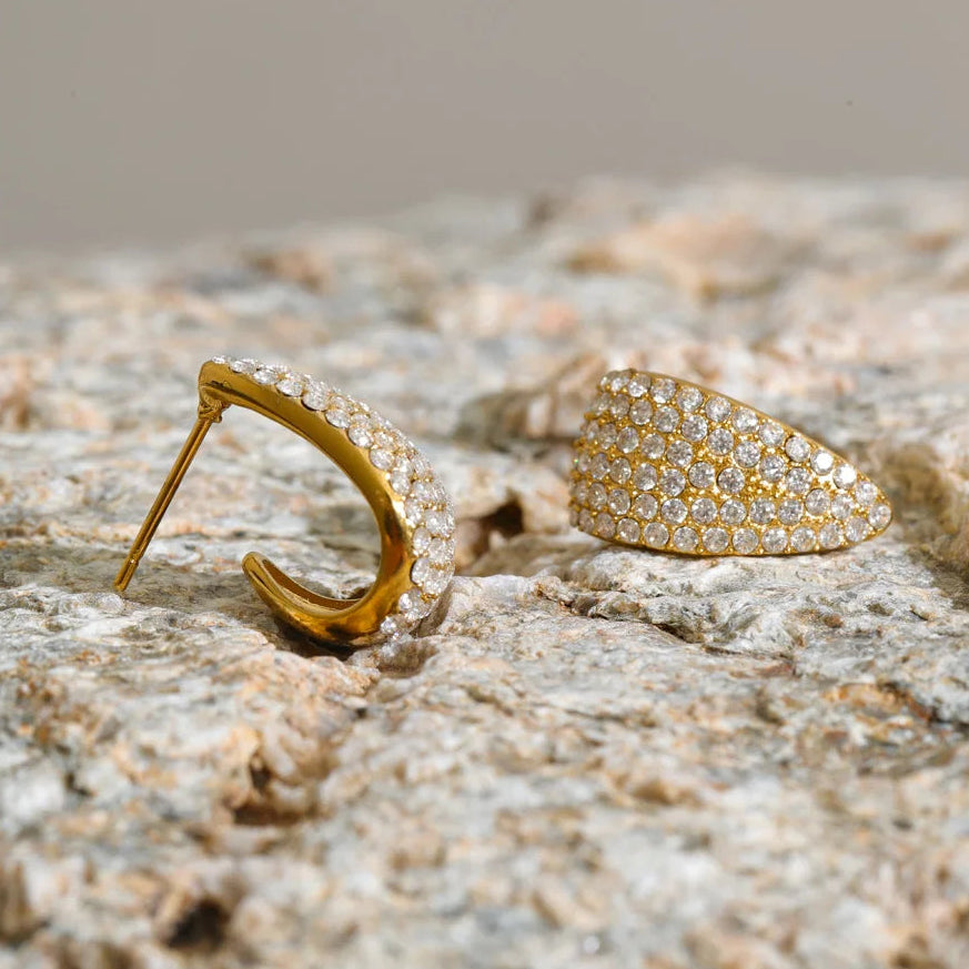 A pair of gold-toned statement earrings, one crescent-shaped and one teardrop-shaped, both heavily embellished with small clear pavé stones, resting on a textured stone surface.
