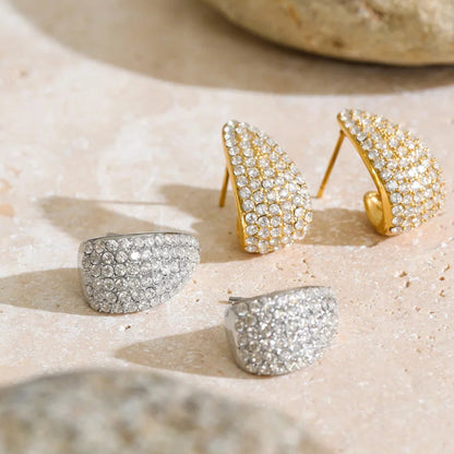 Three pavé hoop earrings displayed on a light stone surface, including two gold-tone and one silver-tone pair, with blurred rocks in the background.