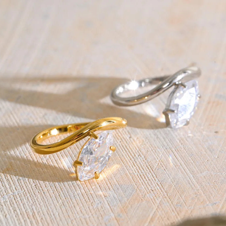 Two small rings with marquise-cut clear stones, one in yellow gold and one in silver, displayed on a textured beige surface with soft shadows.