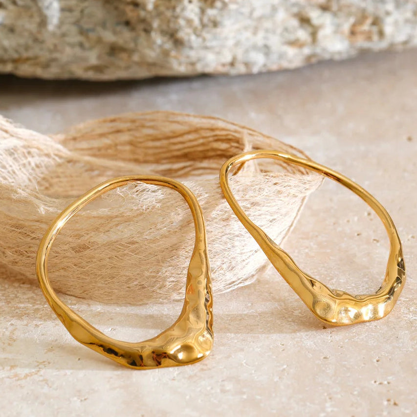 A pair of gold-toned abstract hoop earrings with a hammered texture, displayed on a beige stone surface next to a textured beige fabric and a blurred rock in the background.
