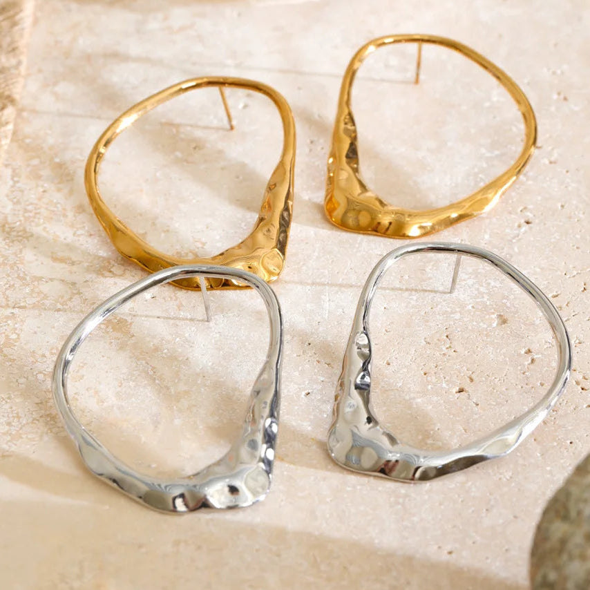 Two abstract open hoop earrings with a textured organic shape, shown in gold and silver finishes on a light beige stone surface.