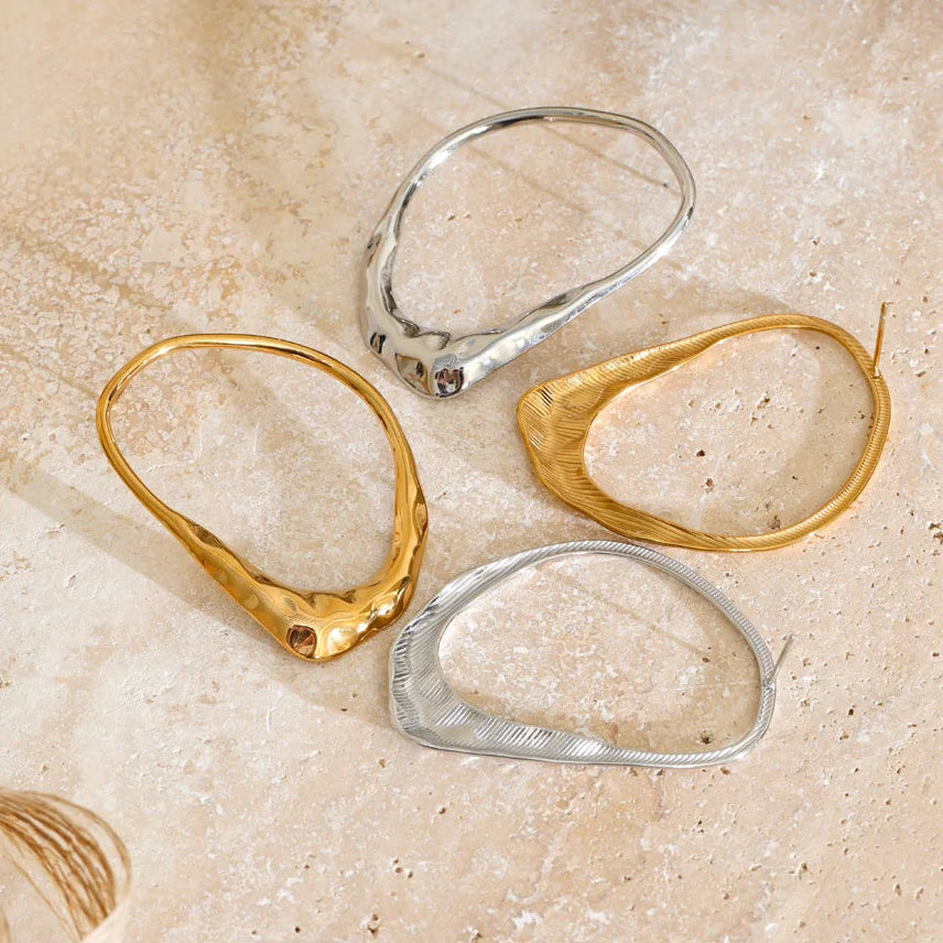 Stainless Steel organic-shaped hoop earrings in textured gold and polished silver, arranged on a light beige stone surface.