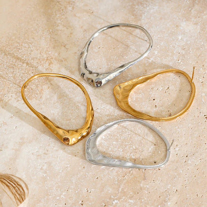 Stainless Steel organic-shaped hoop earrings in textured gold and polished silver, arranged on a light beige stone surface.
