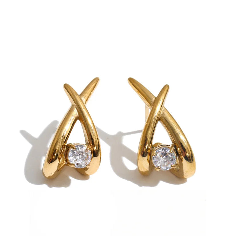 A pair of gold crescent-shaped earrings with a clear heart-shaped gemstone at the base, displayed on a white background with a black script logo below.