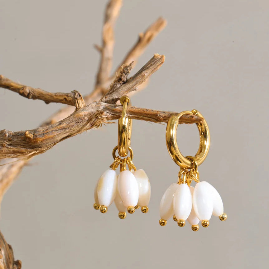 A pair of gold hoop earrings with dangling white pearl-like beads, hanging from a decorative branch against a plain light gray background.