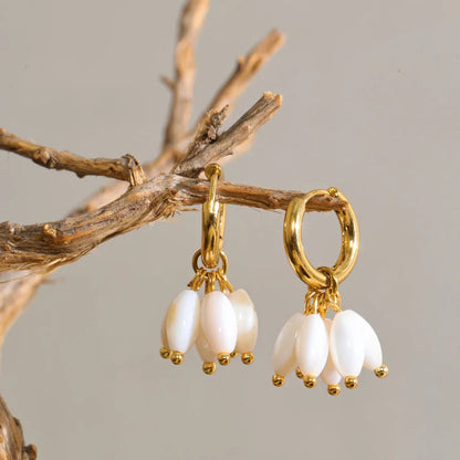 A pair of gold hoop earrings with dangling white pearl-like beads, hanging from a decorative branch against a plain light gray background.
