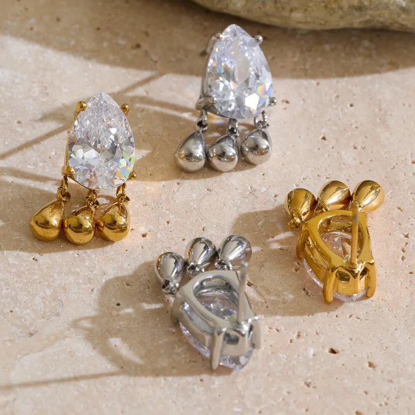 Four clear teardrop-shaped gemstone stud earrings with dangling metal charms, shown in both gold and silver metal finishes on a beige stone-textured surface.
