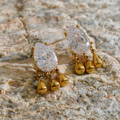 A pair of gold drop earrings with teardrop-shaped clear crystals, displayed on a textured beige stone surface.