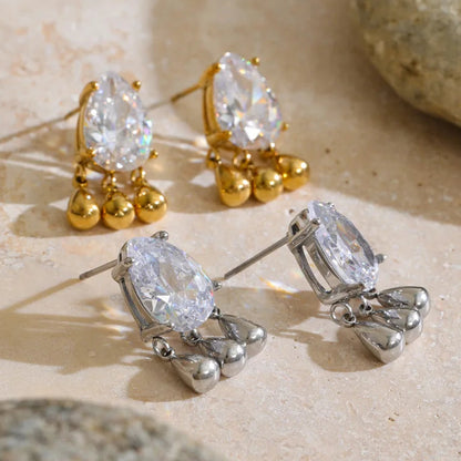 Two pairs of dangling stud earrings displayed on a light beige stone surface. One pair is gold-tone with clear teardrop-shaped stones and small gold bead drops; the other pair is silver-tone with matching clear teardrop stones and silver bead drops.