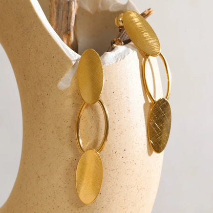 A pair of gold-toned dangle earrings with oval and circular metal elements, displayed on a beige ceramic vase with a piece of driftwood.