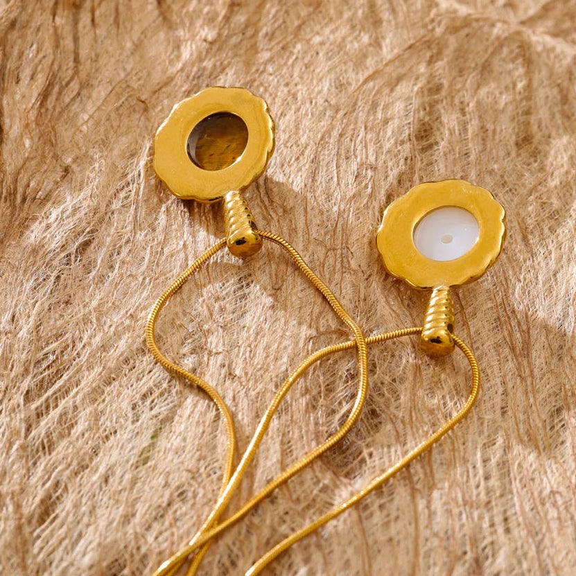 A pair of gold-toned dangle earrings with scalloped circular disc tops, one featuring a dark brown stone and the other a smooth white stone, displayed on a textured beige woven fabric background.
