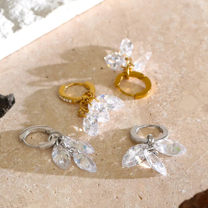 Three pairs of dangling hoop earrings with clear crystal leaf clusters are arranged on a beige stone surface. Two pairs are gold-tone and one pair is silver.