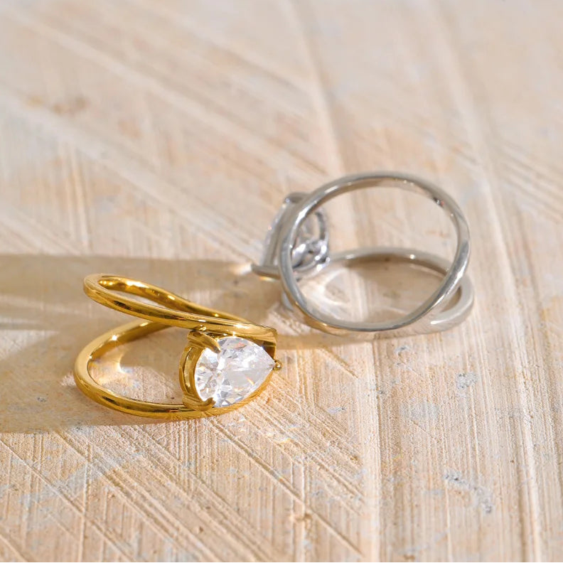 Two minimalist rings on a light wood surface: one gold ring with a clear oval gemstone and a smaller silver ring with a clear oval gemstone. - stainless steel