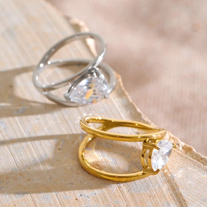 Two rings displayed on a textured beige stone surface: a gold ring with a marquise-cut clear gemstone and a silver ring with a pear-cut clear gemstone.