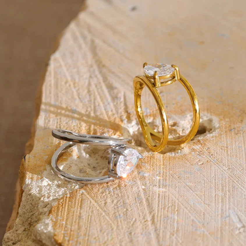 Two rings displayed on a textured stone surface: one gold ring with a clear rectangular gemstone and one silver ring with a clear marquise-shaped gemstone.