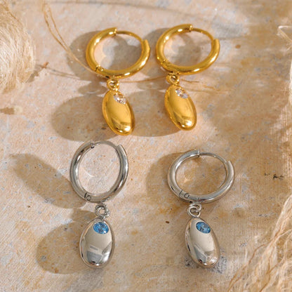 Two pairs of drop hoop earrings displayed on a textured beige surface. The top pair features gold-tone hoops with polished gold teardrop charms and a small clear stone.