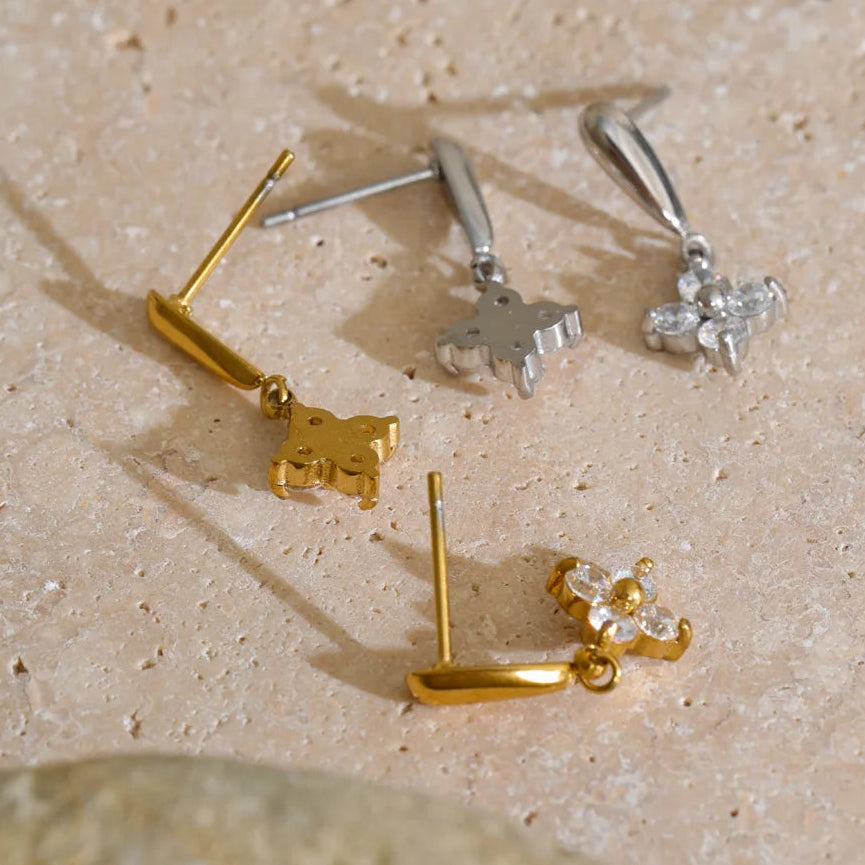 Four small stud earrings arranged on a beige stone surface: two gold-tone earrings with animal-shaped charms and two silver-tone earrings with floral/crystal charms.