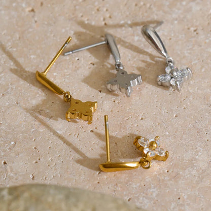 Four small stud earrings arranged on a beige stone surface: two gold-tone earrings with animal-shaped charms and two silver-tone earrings with floral/crystal charms.