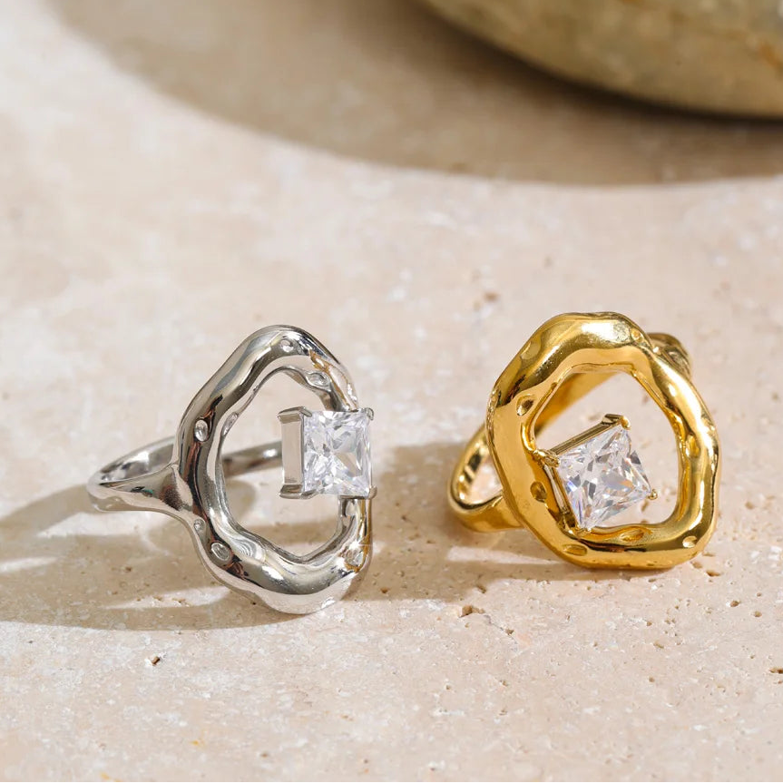 Two abstract openwork rings with square clear gemstones, one in silver-tone metal and one in yellow-gold metal, displayed on a light beige stone surface.