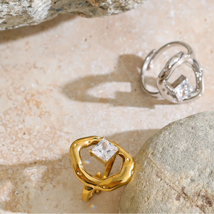 Two statement rings with square clear gemstones displayed on a textured beige stone surface with rocks. One ring is gold-toned and the other is silver-toned.