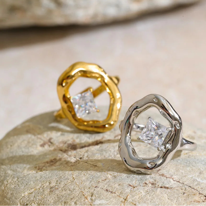 Two textured ring earrings with bezel-set clear square gemstones, one in yellow gold and one in silver, displayed on a natural stone surface. Stainless Steel