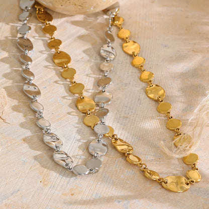 Two beaded necklaces with gold and silver disc-like beads, arranged on a textured beige fabric next to a decorative shell.