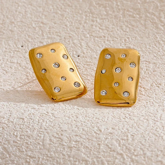 A pair of gold rectangular stud earrings with small clear rhinestones, displayed on a textured beige surface next to decorative stones. Waterproof stainless steel.