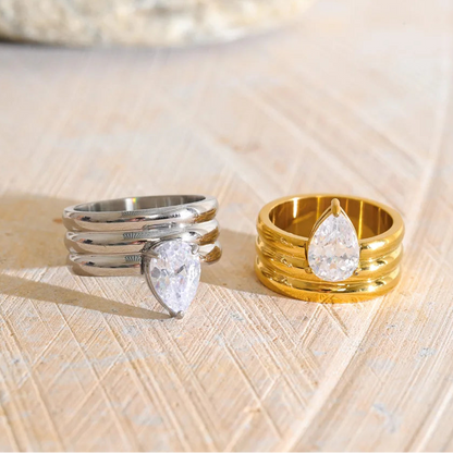 Two pear-cut gemstone rings displayed on a light wood surface. One ring is silver-toned with three stacked bands and a pear-shaped clear stone, and the other is gold-toned with three stacked bands and a matching pear-shaped clear stone.