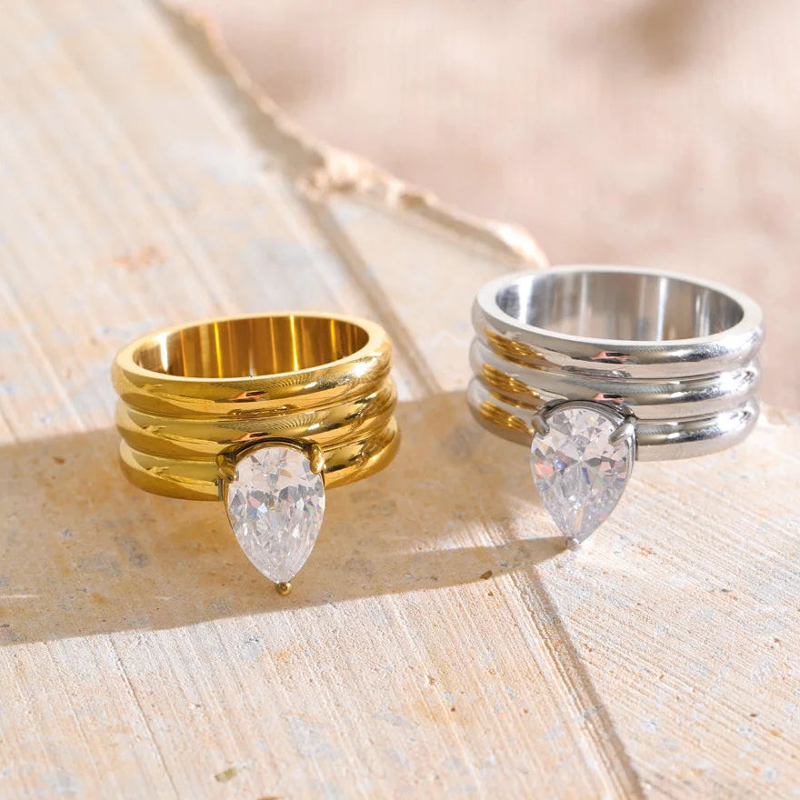 Two stacked ring sets with pear-cut clear gemstones, one in polished yellow gold and one in polished silver, displayed on a light wooden surface.