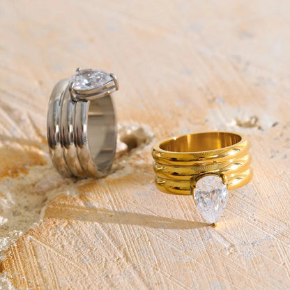 Two stacked ring sets displayed on a textured beige stone surface. One set is silver-toned with a pear-cut clear gemstone, and the other is gold-toned with a pear-cut clear gemstone.