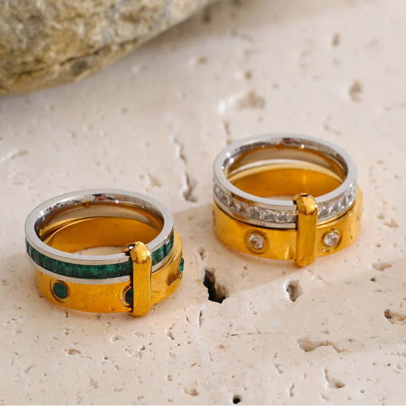Two gold and silver rings with green and white accents, displayed on a textured beige stone surface with a blurred rock in the background.