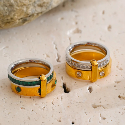 Two gold and silver rings with green and white accents, displayed on a textured beige stone surface with a blurred rock in the background.