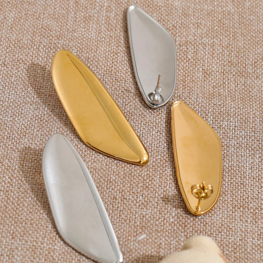 Four minimalist leaf-shaped earrings in polished gold and silver finishes, arranged on a textured beige linen background.