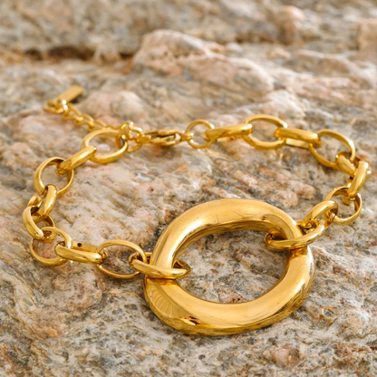 A gold chain bracelet with a large textured circular ring pendant, displayed on a rough stone surface.