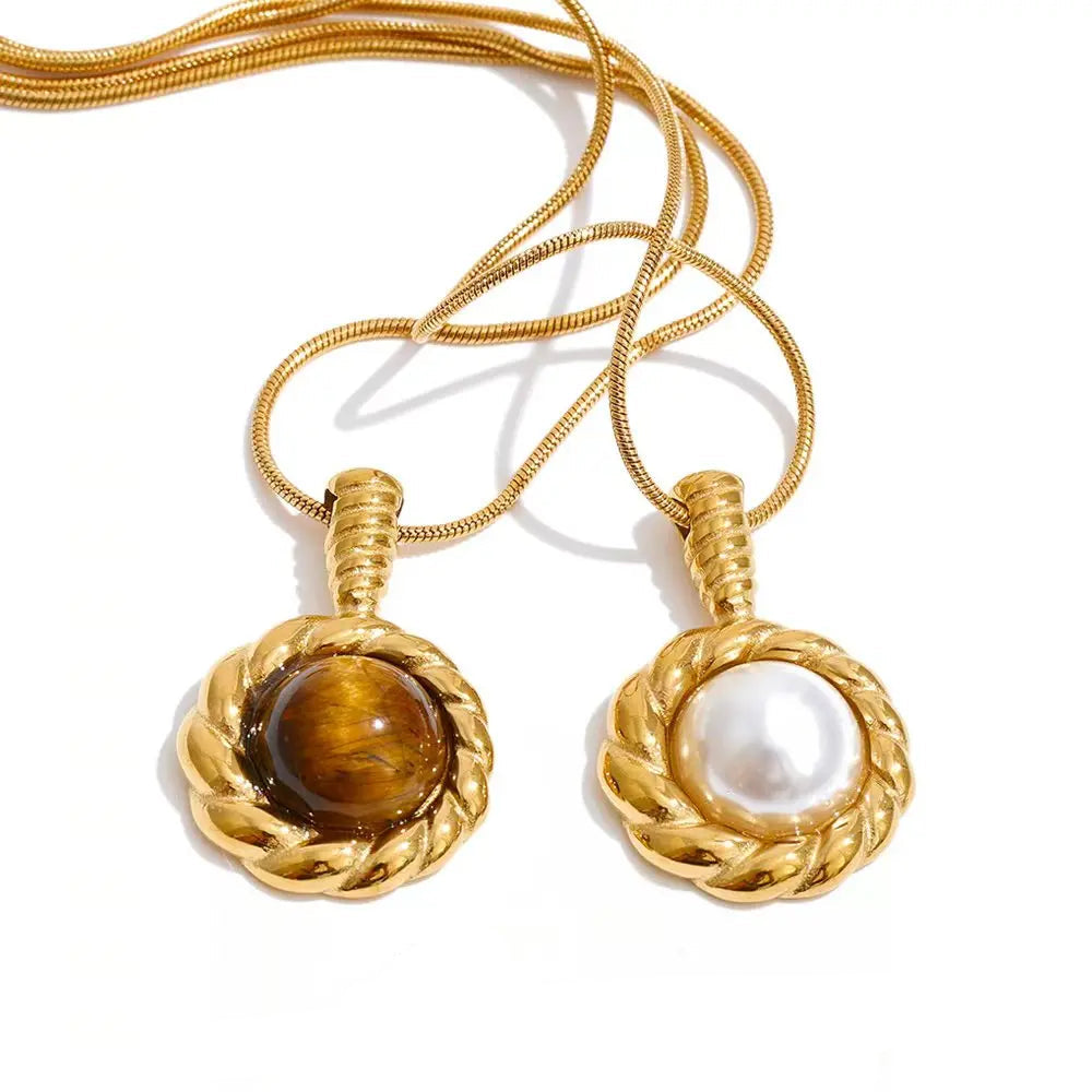 Two gold pendant necklaces on a white background, one with a round brown tiger's eye stone and the other with a white pearl, both set in a twisted rope-style gold bezel. 
