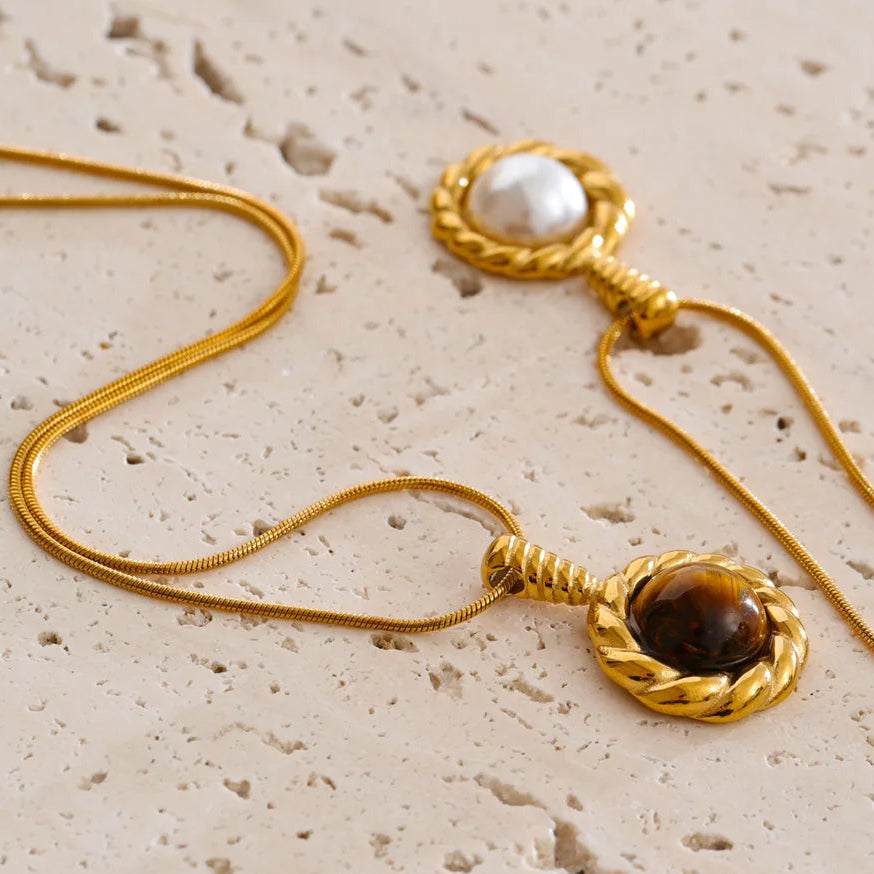 Two gold-toned snake chain necklaces with round pendant settings, one featuring a white pearl and the other a brown tiger's eye stone, displayed on a textured beige stone surface.