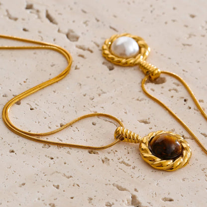 Two gold-toned snake chain necklaces with round pendant settings, one featuring a white pearl and the other a brown tiger's eye stone, displayed on a textured beige stone surface.