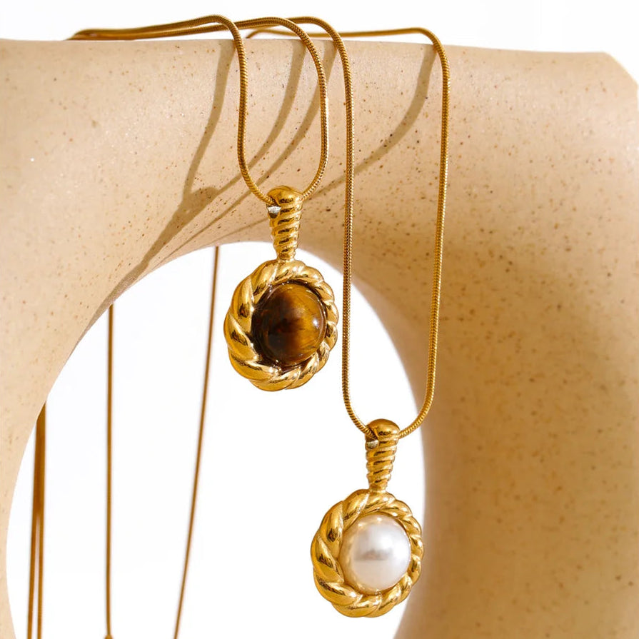 Two gold pendant necklaces draped over a beige jewelry display bust. One pendant features a brown tiger's eye stone, and the other features a white pearl, both set in twisted gold bezels on fine gold chains.