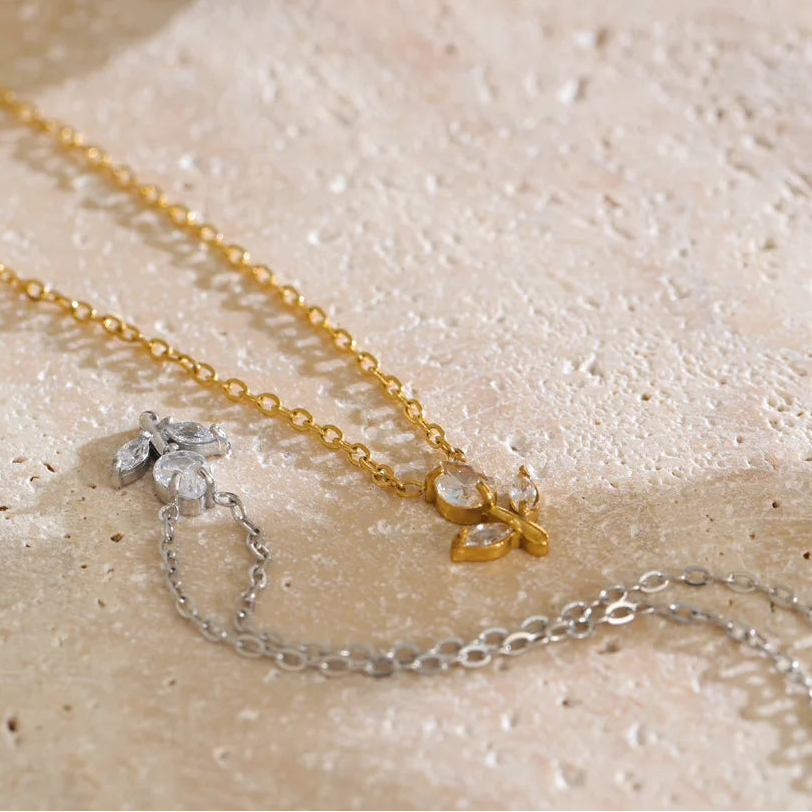 A delicate gold chain necklace with a small gold leaf pendant and a silver chain with a silver leaf pendant, both adorned with clear crystals, displayed on a textured beige stone surface.