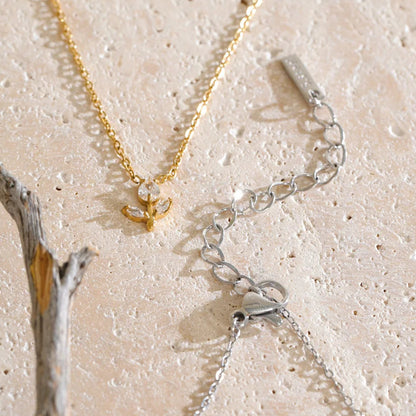 Two delicate necklaces laid flat on a light beige textured surface: one gold chain with a small gold pendant and one silver chain with a silver pendant, both shown with extender links. A small piece of driftwood is visible at the left edge.