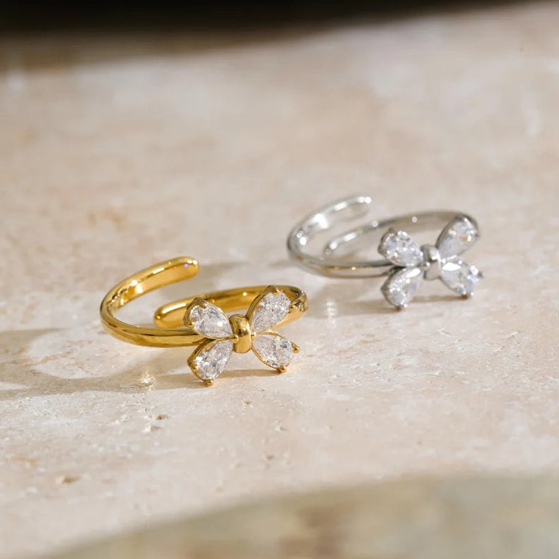 Two butterfly-shaped open rings, one in yellow gold and one in silver, both adorned with clear crystal-like butterfly wings, displayed on a beige stone surface.