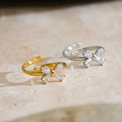 Two butterfly-shaped open rings, one in yellow gold and one in silver, both adorned with clear crystal-like butterfly wings, displayed on a beige stone surface.